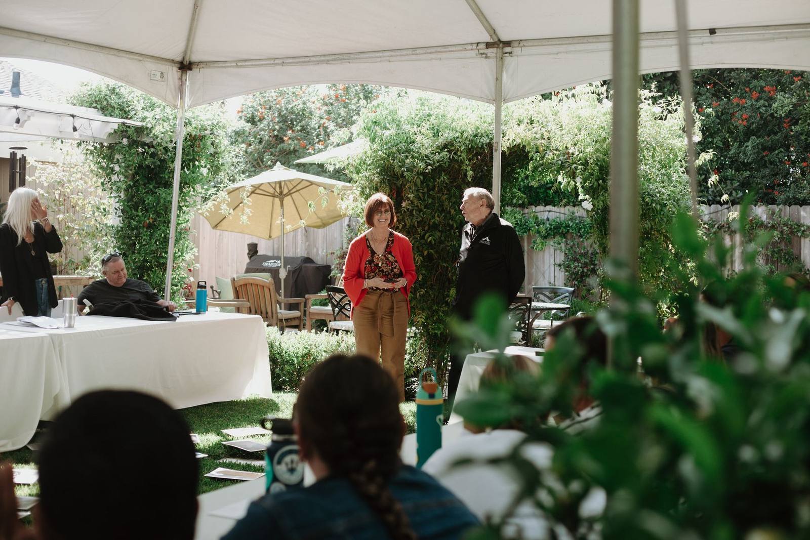 A cohort gathered under a tent in a garden while two leaders speak
